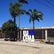 Fachada frontal da escola, um prédio de um andar com tijolos à vista. O nome da escola está pintado em azul na entrada. Veem-se três palmeiras e uma árvore de menor porte. Ainda, em frente à cerca branca, há um letreiro que diz "Eu amo CEMA" e um banco.