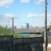 Em primeiro plano, o muro de contenção da escola, com uma parte com cerca e outra sem. Atrás do muro, um campo de capoeira alta. Ao lado do muro vê-se um poste e, ao fundo, uma casa azul.