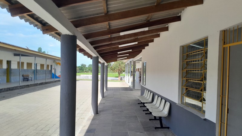 Vê-se o pátio da escola, com um corredor coberto à frente e um dos prédios à direita e outro à esquerda.